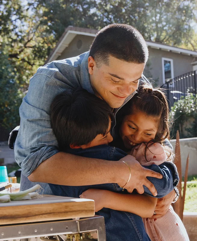 A man with short dark hair smiles and leans over to hug two kids while standing outside in the yard.