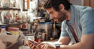 A white bearded man is looking phone while leaning on a workshop desk.