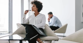 A young Black woman in a white blouse looking contemplative sits in a light-filled office with a portfolio on her lap. 