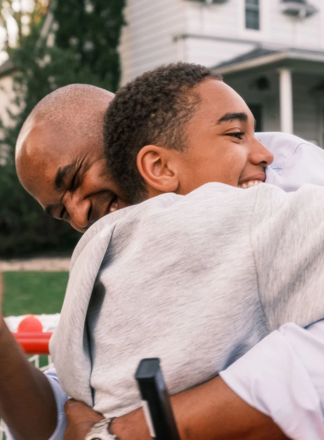 A Black father and son embrace in their front yard, their white house in the background.