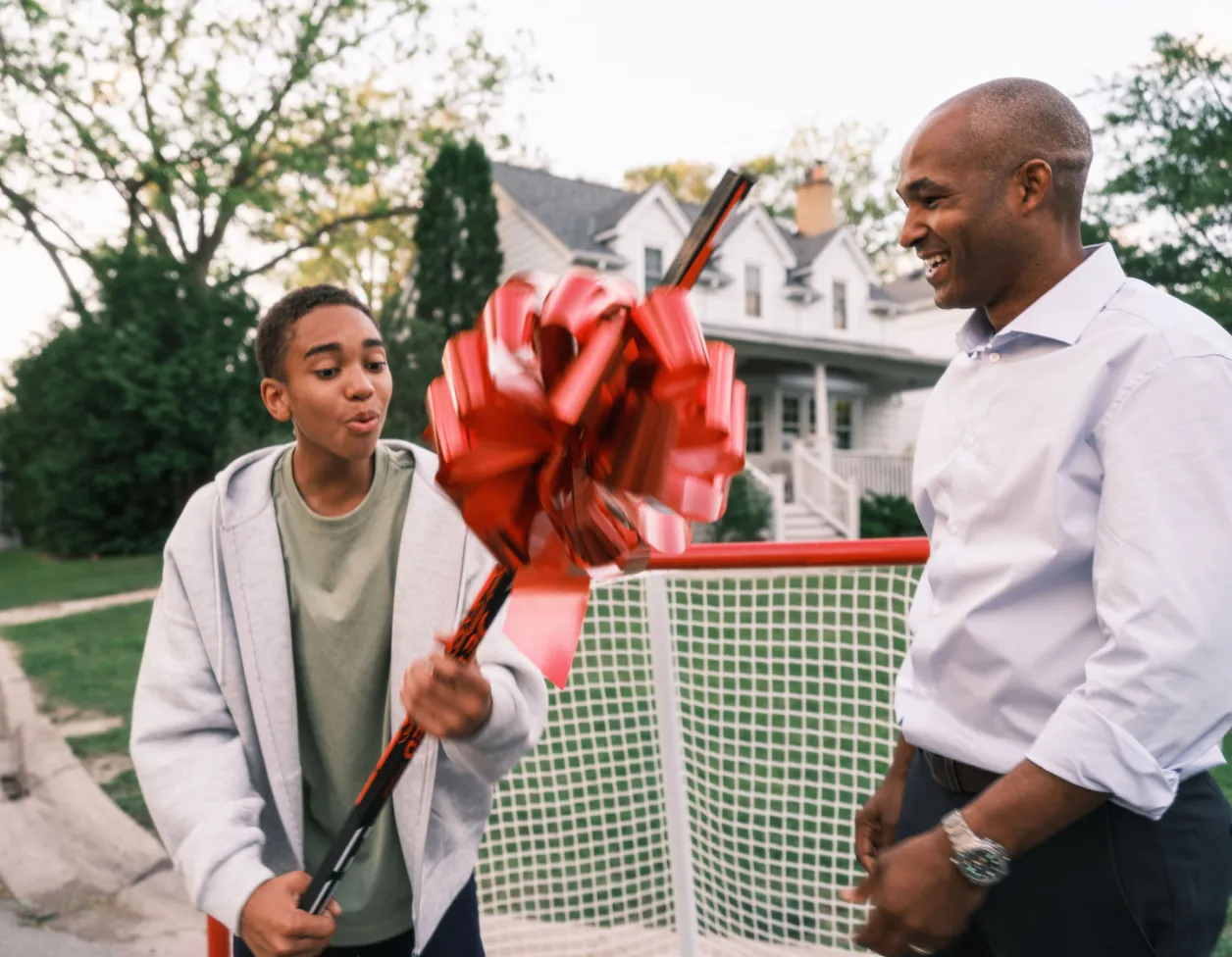 A Black teenage boy holds a new hockey stick with a big red bow while his father looks on proudly.