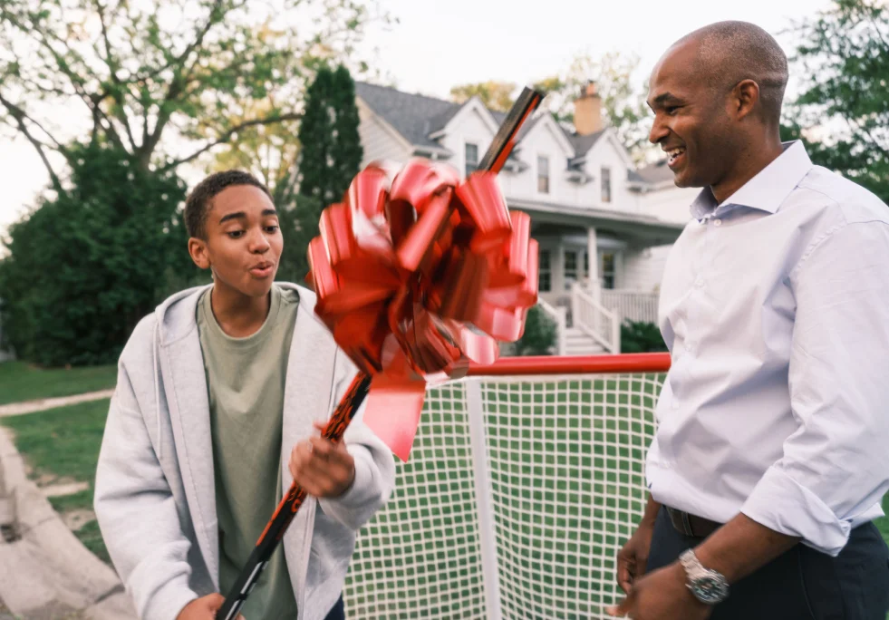 A Black teenage boy holds a new hockey stick with a big red bow while his father looks on proudly.