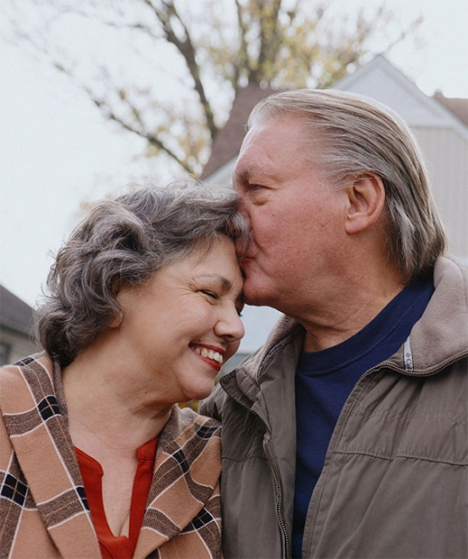 A mature white man plants a loving kiss on his wife's forehead.