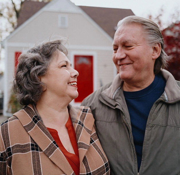 A mature white couple smile while exchanging a loving look outside their home.