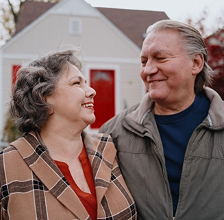 A mature white couple smile while exchanging a loving look outside their home.