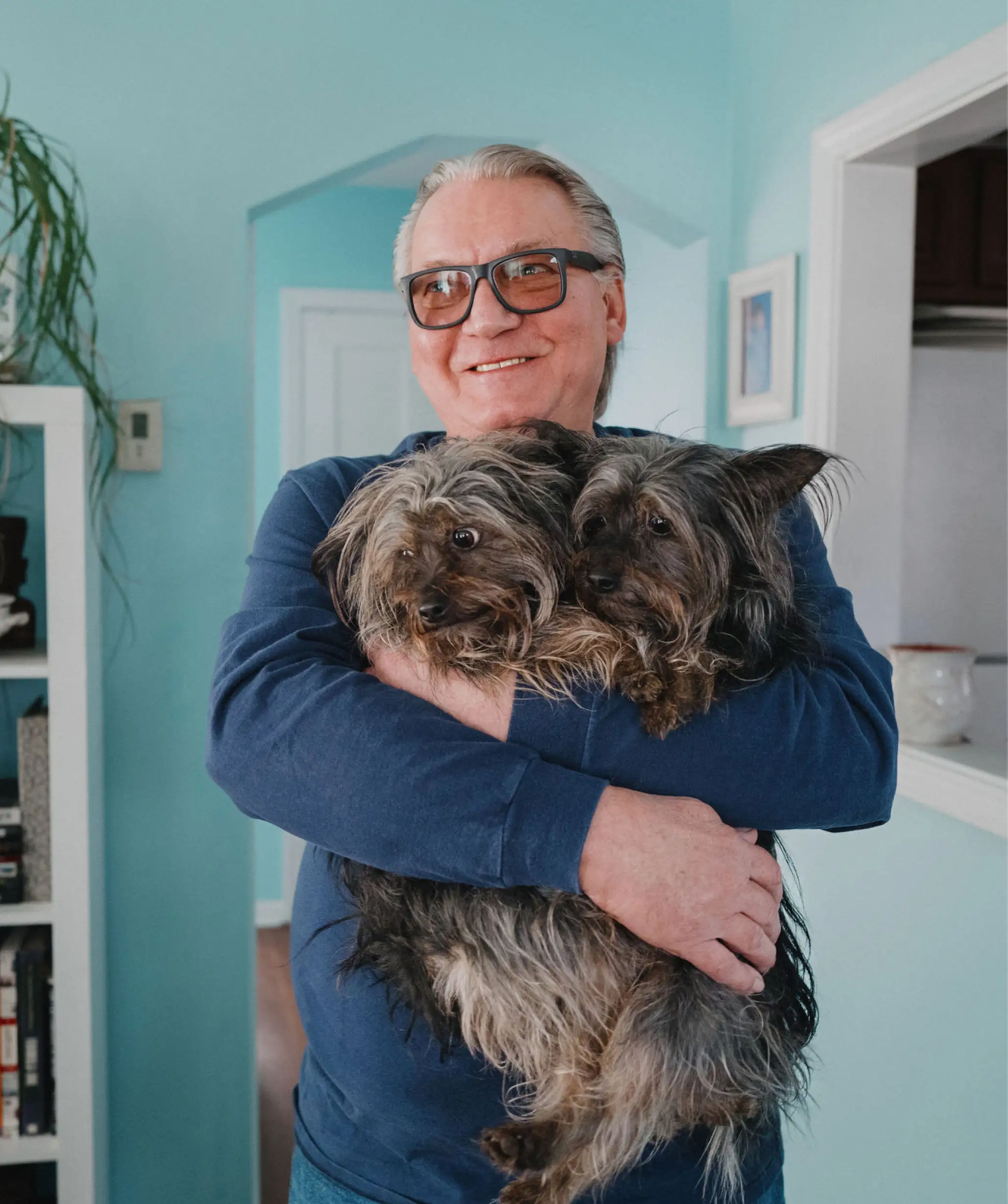 A mature man wearing glasses holds his two Yorkshire terriers inside his home.