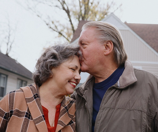 A mature white man plants a loving kiss on his wife's forehead.