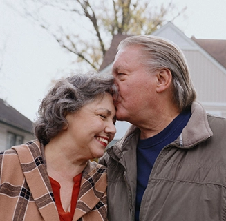 A mature white man plants a loving kiss on his wife's forehead.