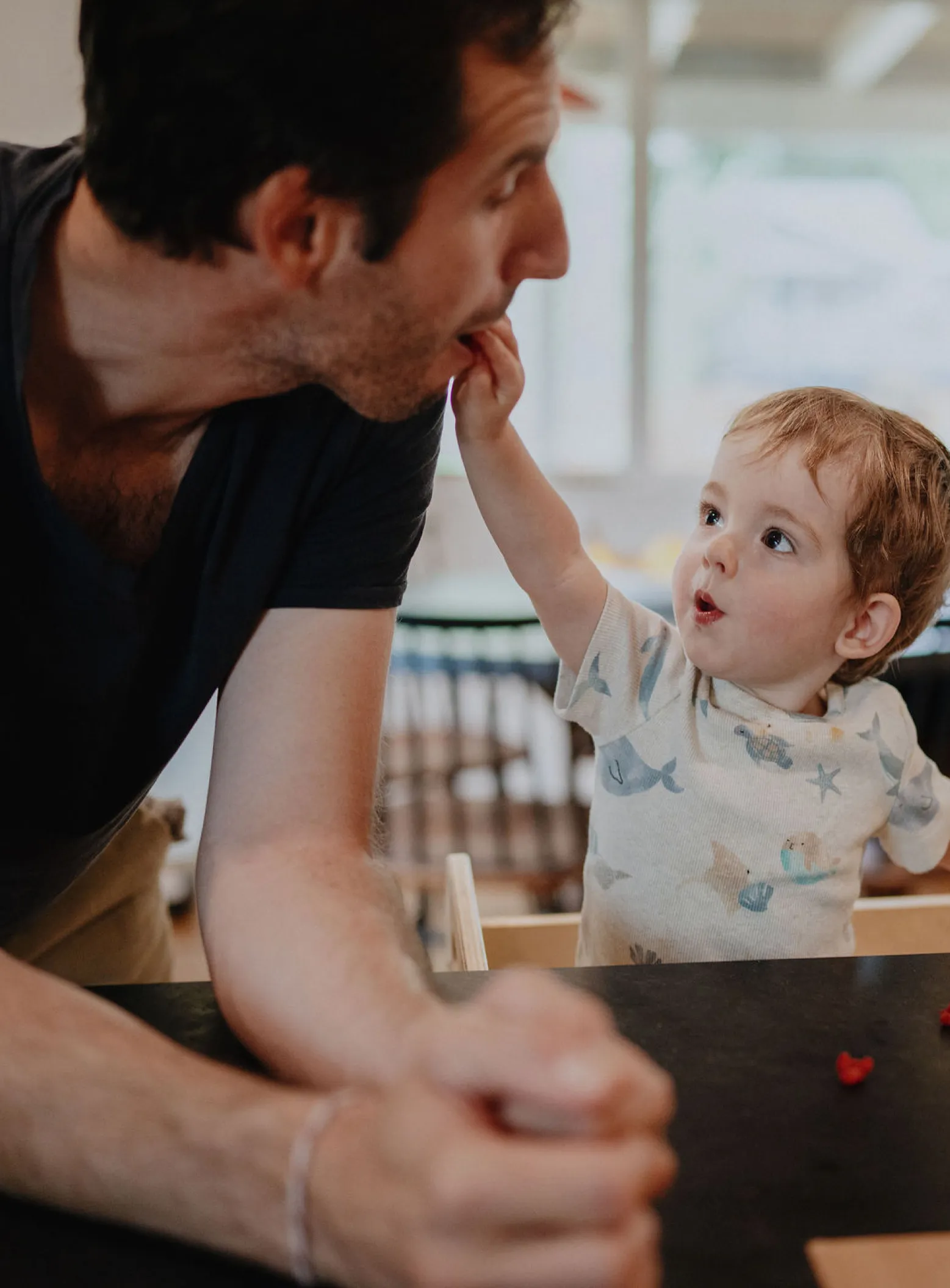 Un niño rubio pone un pequeño refrigerio en la boca de su padre mientras este se inclina sobre la mesa del comedor.