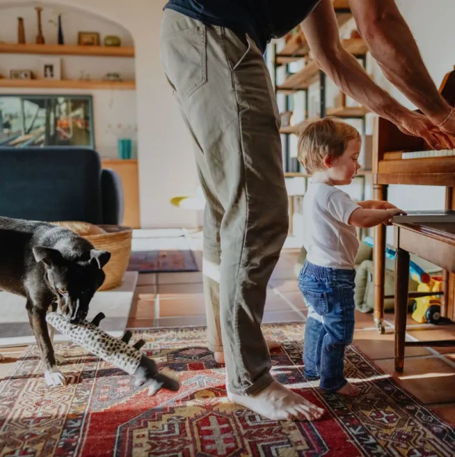 A barefoot toddler and his dad stand in front of a piano while the father plays and the family dog chews on a toy behind them.