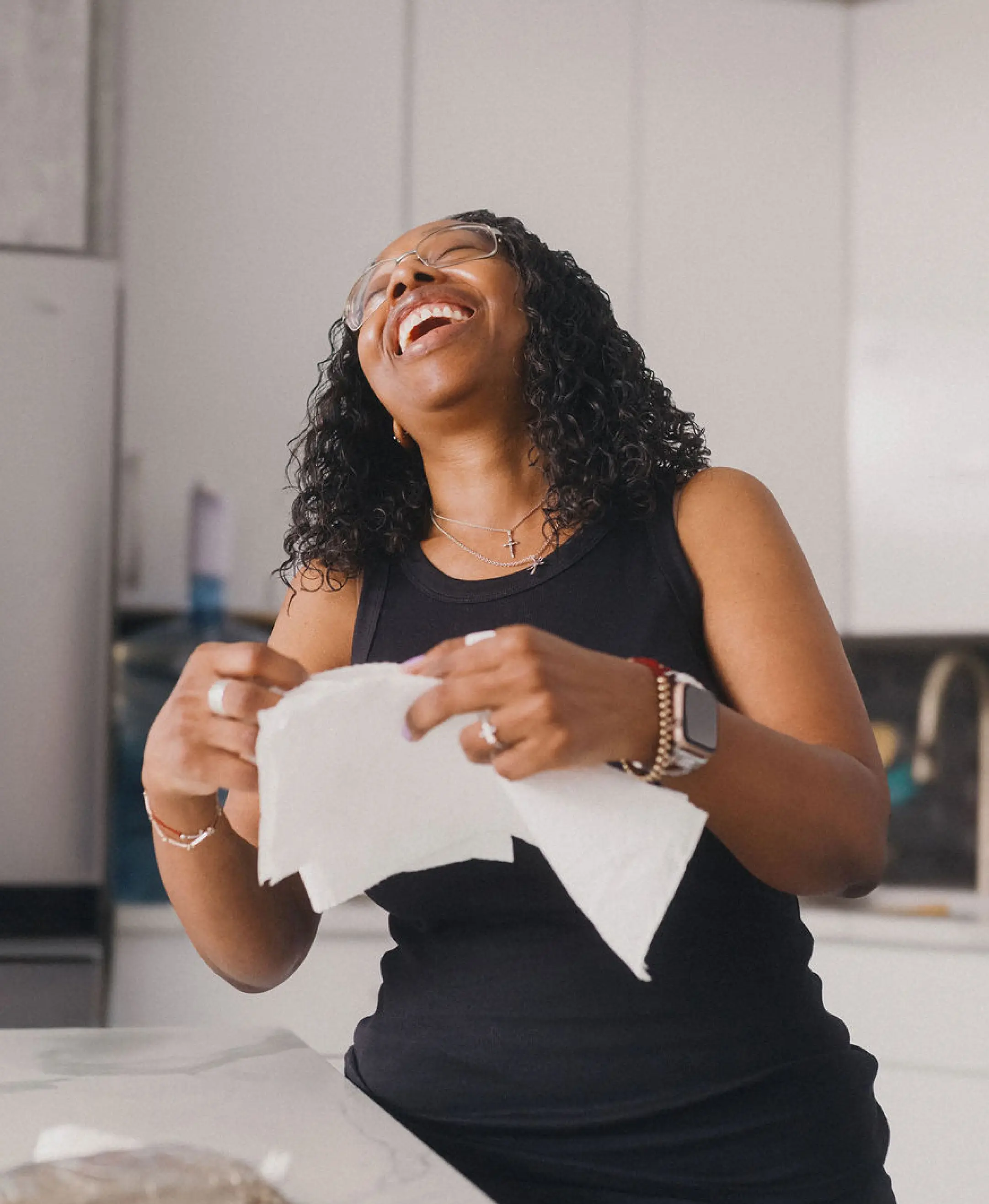 A Black woman in her 30s wearing a black sleeveless shirt and holding a piece of paper towel enjoys a robust laugh in her kitchen.