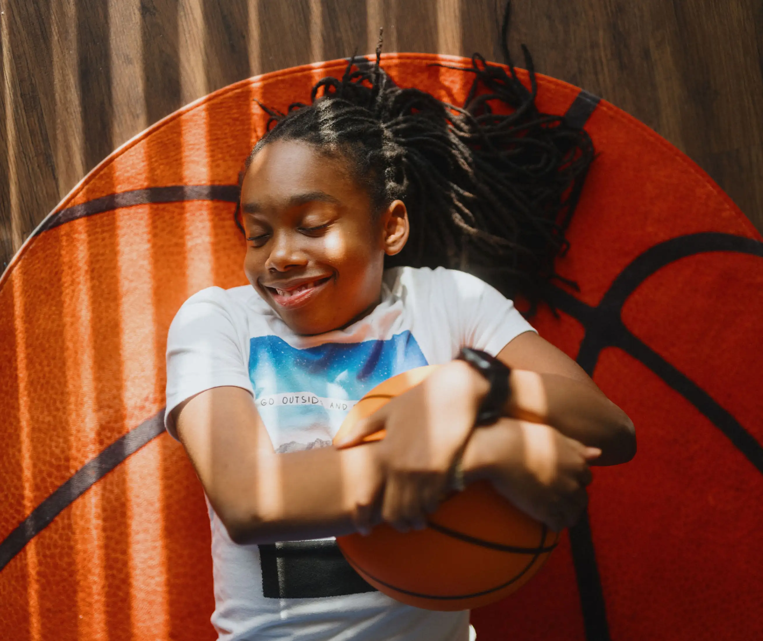 A young Black girl lays on a gym floor while hugging a basketball.