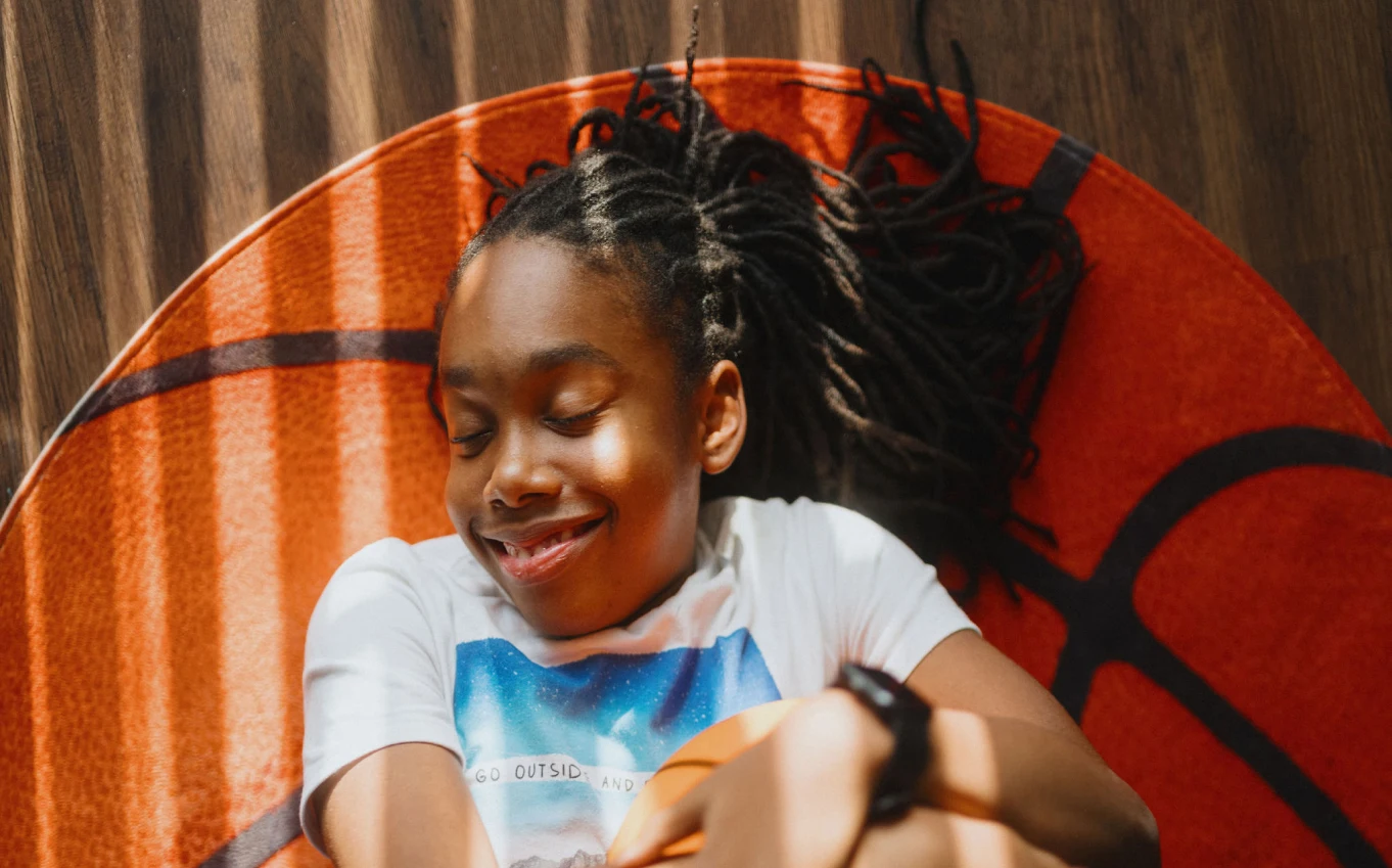 A young Black girl lays on a gym floor while hugging a basketball.