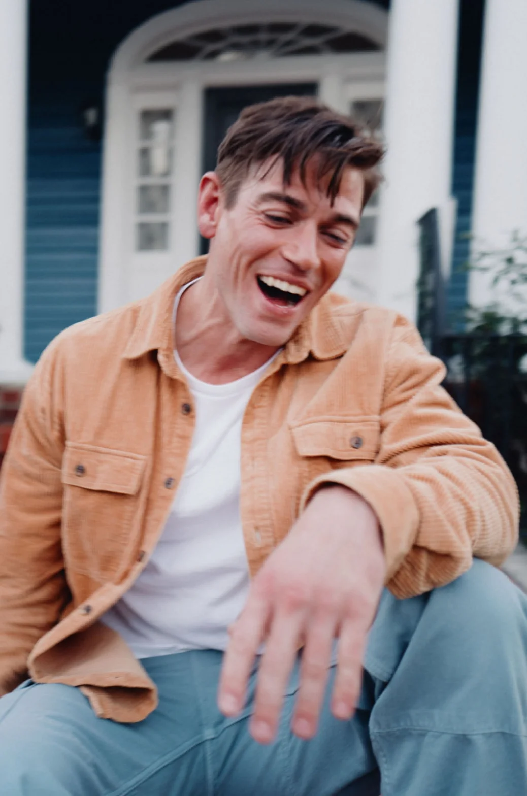 A joyful white man wearing a white shirt and tan jacket sits on the steps in front of his blue house.