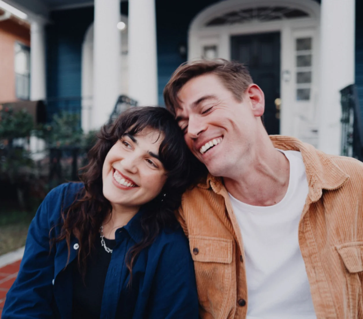 A smiling dark-haired woman rests her head on a brown-haired man's shoulder while they sit in front of their blue house.