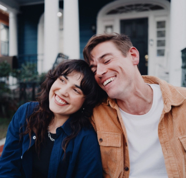 A smiling dark-haired woman rests her head on a brown-haired man's shoulder while they sit in front of their blue house.