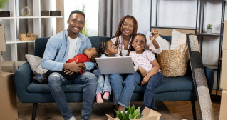 A Black couple with three young children celebrate getting the keys to their new home while sitting on the couch together.