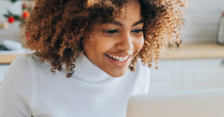 A Black woman wearing a white turtleneck shirt smiles while looking at a laptop in her kitchen.