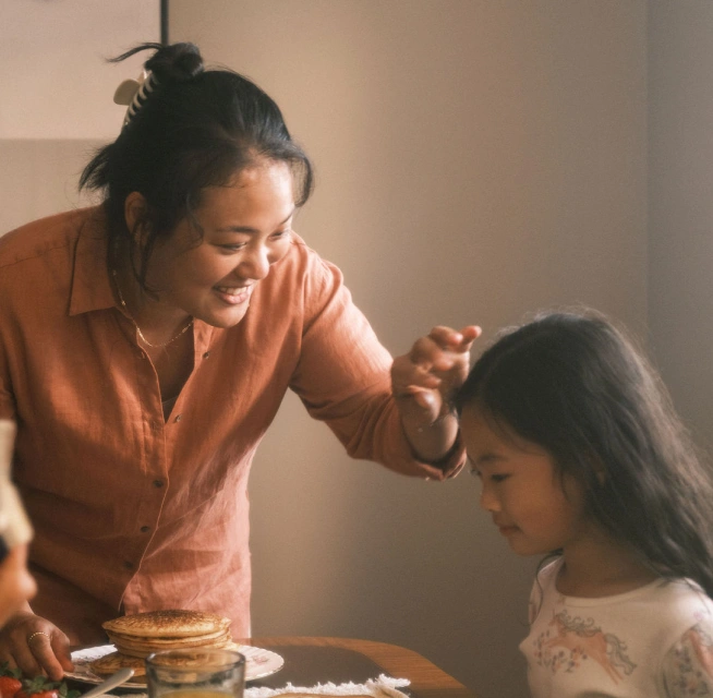 Una joven madre asiática se inclina sobre una mesa de comedor, arreglando el cabello de su hija mientras sus hijos están desayunando.