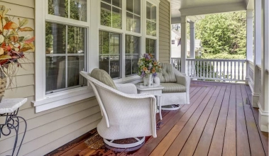  An inviting porch with a brown wood floor, white railings, two white chairs and fall floral arrangements.