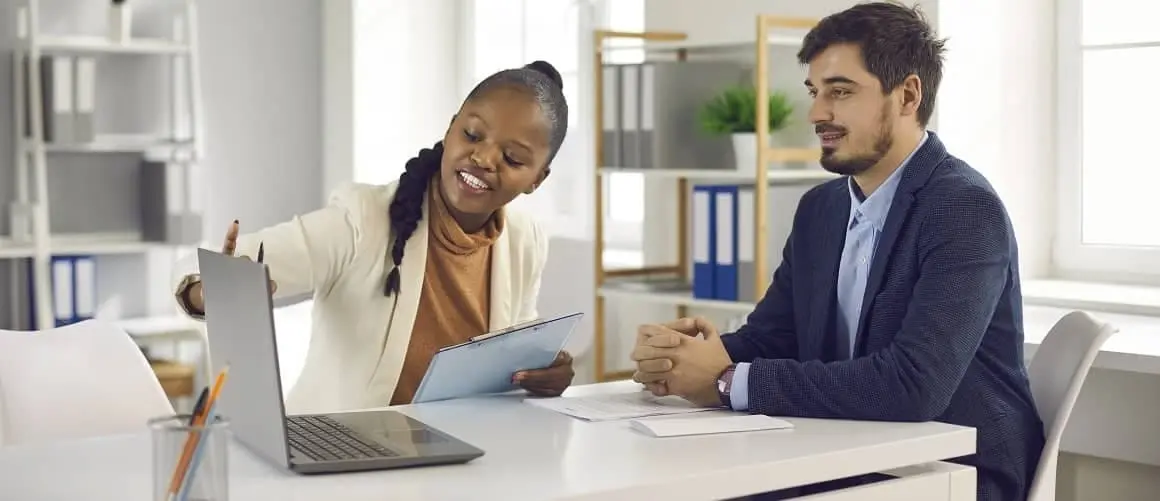 A Black woman in a cream blazer and tan turtleneck explains short term mortgages to a male friend using a laptop.