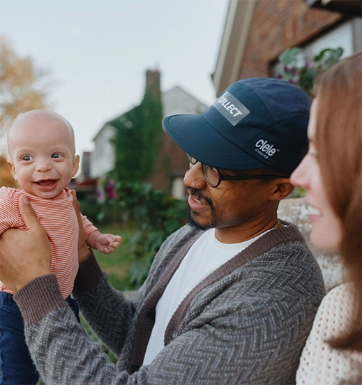 Una familia multiracial se encuentra frente a su casa de ladrillos, el padre sostiene al bebé frente a él mientras la madre sonríe.