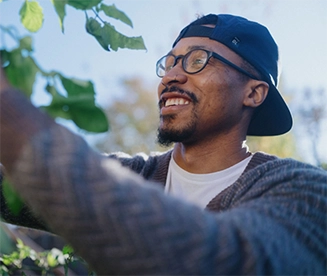 Un hombre negro con gafas, gorra de béisbol al revés y un anillo en la nariz sonríe mientras poda un árbol en su patio trasero.