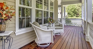 An inviting porch with a brown wood floor, white railings, two white chairs and fall floral arrangements.