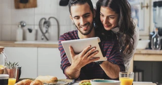 A couple research lowering their mortgage payment on their tablet while eating breakfast in their kitchen.