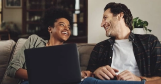 A Black woman in her thirties looks at her partner, a white man in a plaid shirt, balancing a laptop on her leg as they lounge on a couch together. 