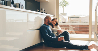 Una pareja blanca sentada en el suelo de la cocina hablando mientras sostienen tazas de café.