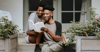 A Black man reads from a book to his partner as they sit on the porch of their home.