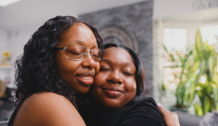 Two Black women share a warm hug in a living room.