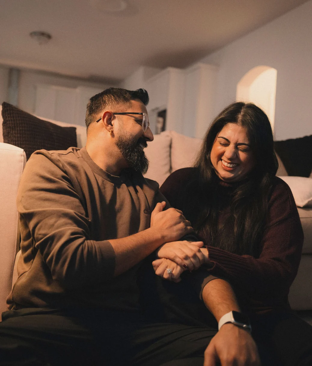 A dark-haired man and woman hug each other's arms while sitting on the floor in front of a white couch in their new home.