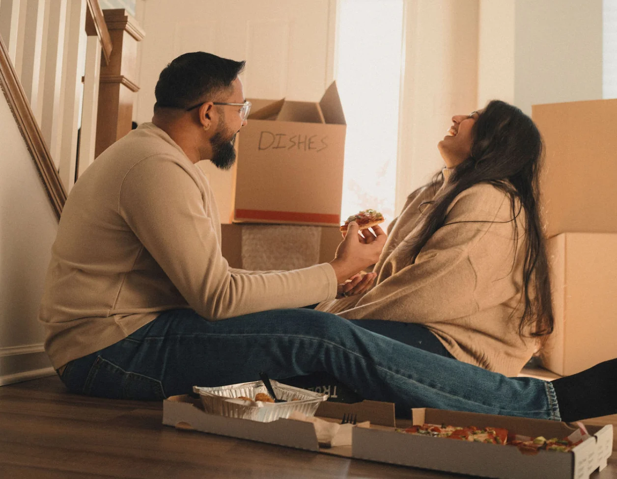 Una pareja de cabello oscuro se sienta en el suelo de su nuevo hogar, rodeada de cajas de mudanza mientras comen pizza y comparten una risa.