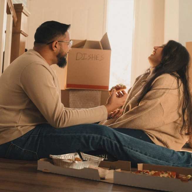 Una pareja de cabello oscuro se sienta en el suelo de su nuevo hogar, rodeada de cajas de mudanza mientras comen pizza y comparten una risa.