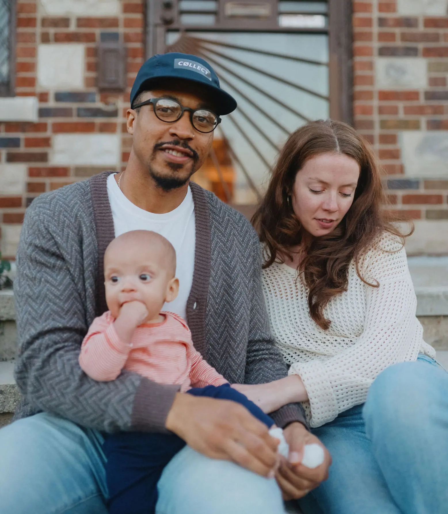 A young couple wearing blue jeans and sweaters sit with their baby on the front porch of their home.