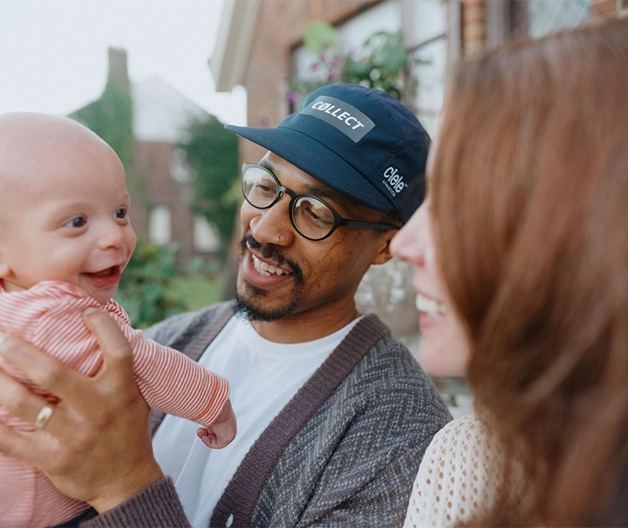 A Black man in a blue baseball cap shows a bald smilling baby in a pink shirt to a white woman with long auburn hair.