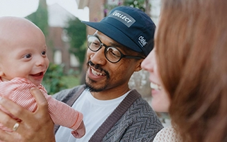 A Black man in a blue baseball cap shows a bald smilling baby in a pink shirt to a white woman with long auburn hair.