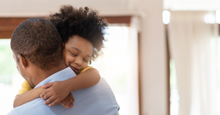 A Black toddler hugs his dad around the neck while his dad stands and holds him.