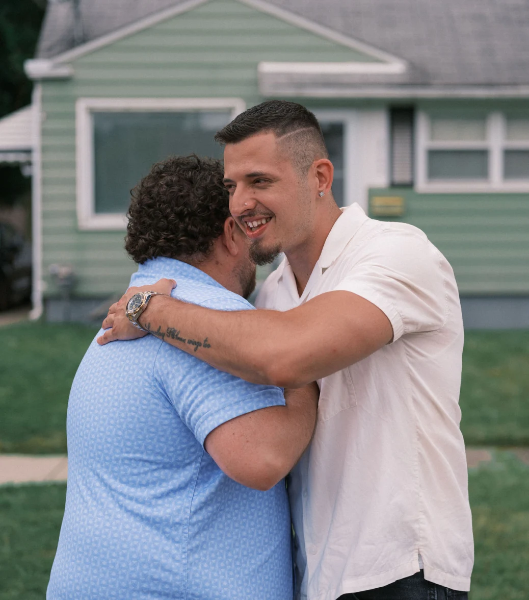 A man with curly brown hair hugs a man with short brown hair in front of a green house with white trim.