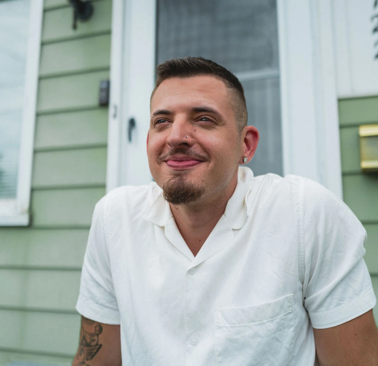 A brown-haired man with a white collared shirt, earrings, and a nose ring sits on the front porch of a green house with white trim.