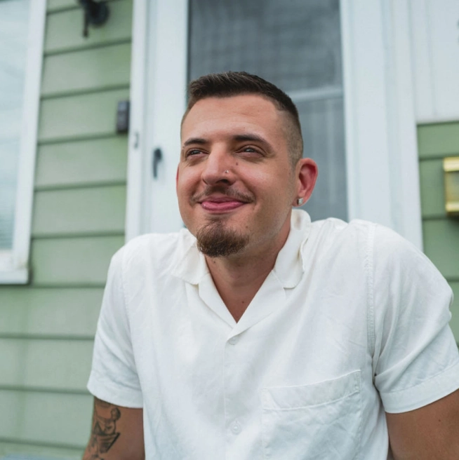 A brown-haired man with a white collared shirt, earrings, and a nose ring sits on the front porch of a green house with white trim.