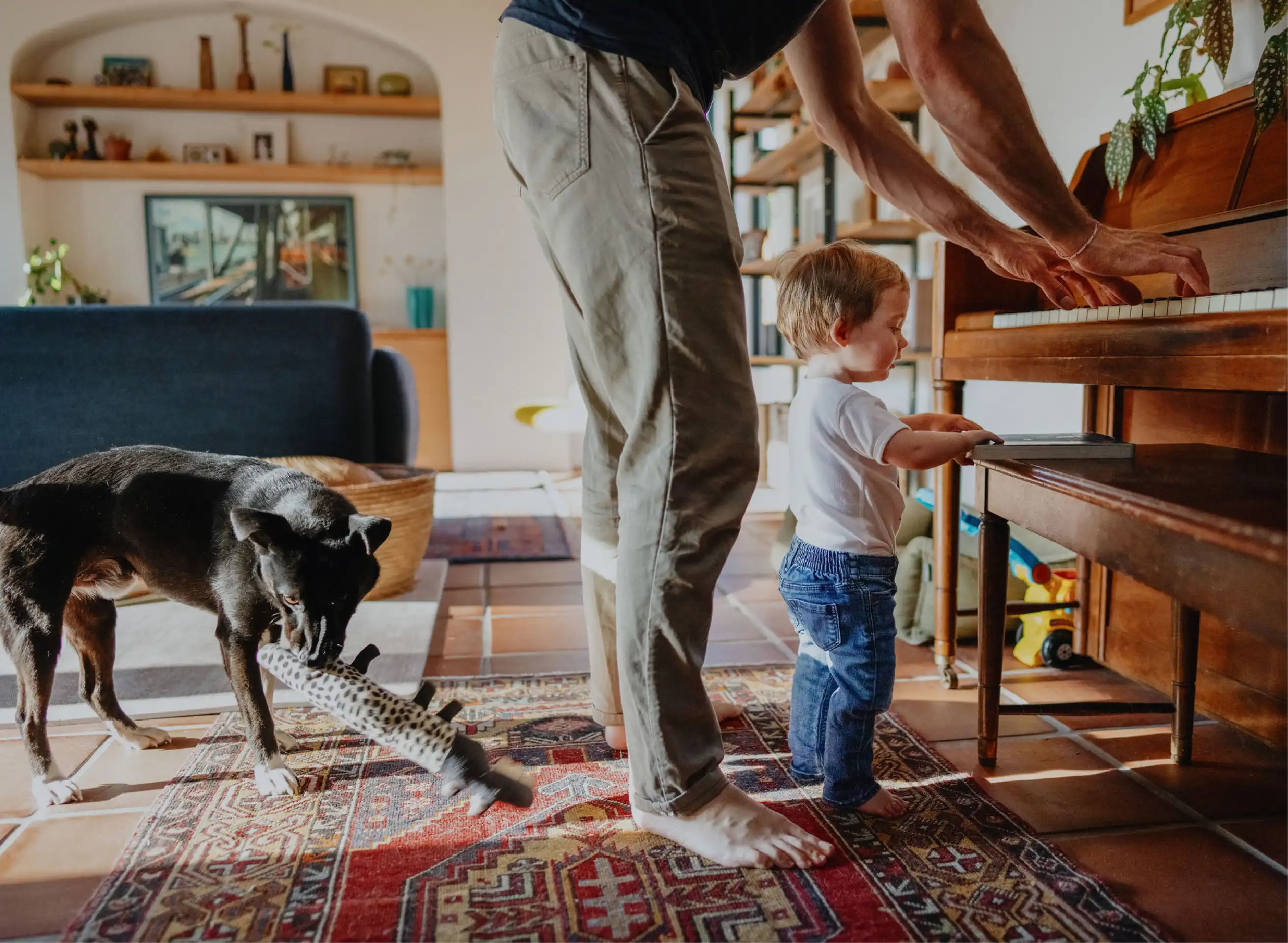 A barefoot toddler and his dad stand in front of a piano while the father plays and the family dog chews on a toy behind them.