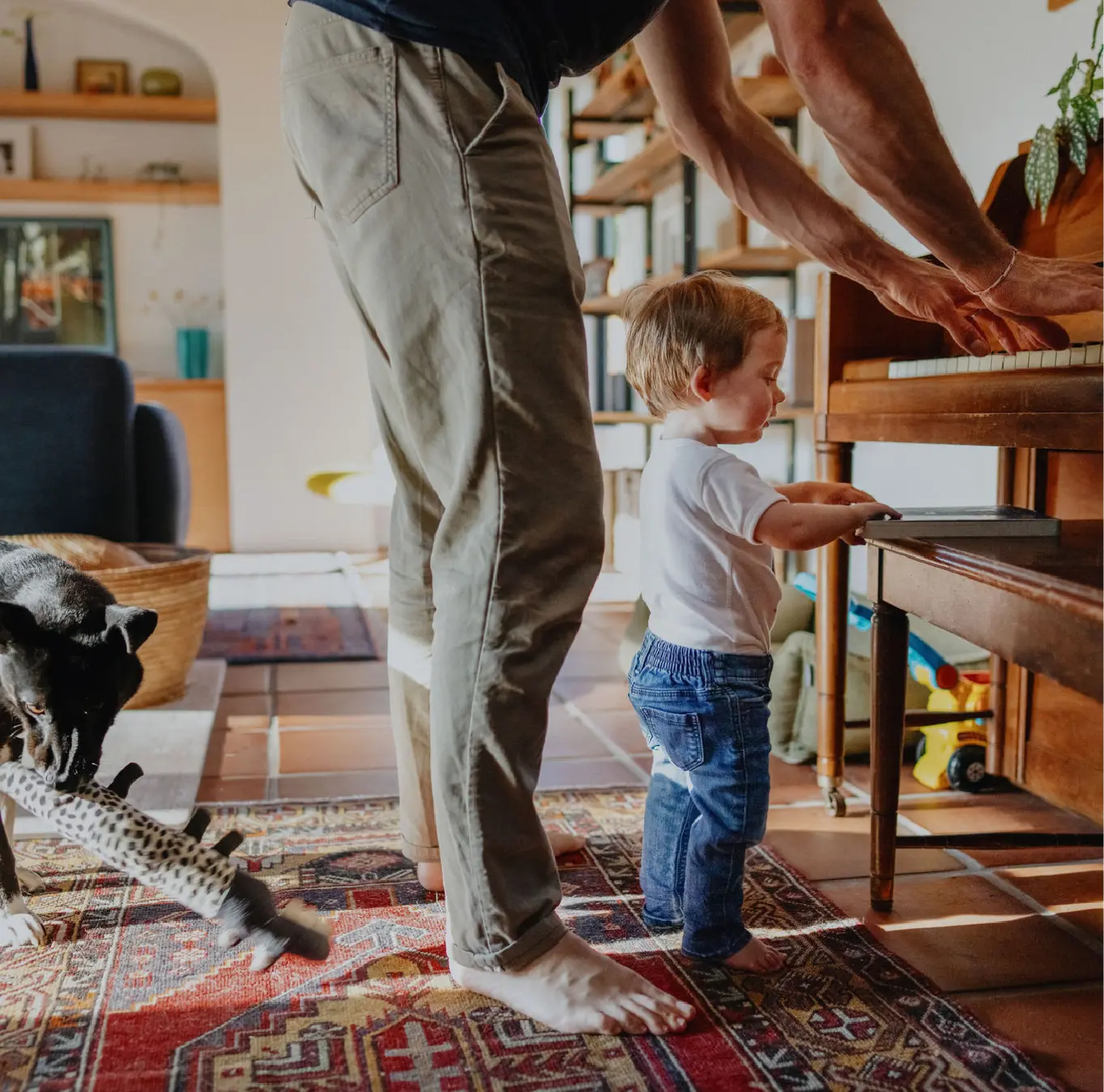 Un niño descalzo y su padre están parados frente a un piano mientras el padre toca y el perro de la familia mastica un juguete detrás de ellos.