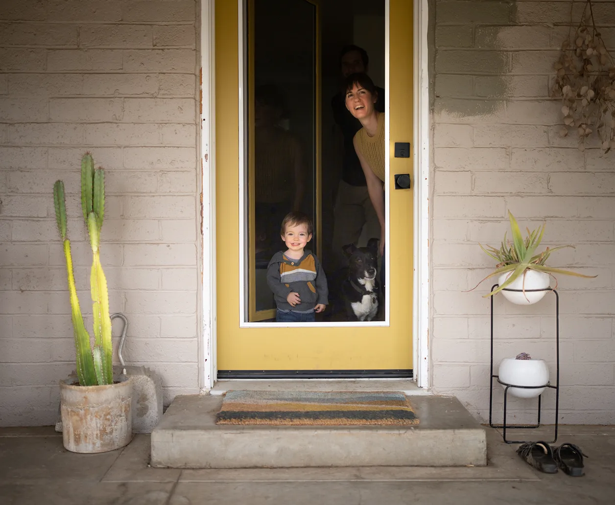 A toddler, a black dog, and the toddler's mom peer through the large window of a yellow front door.