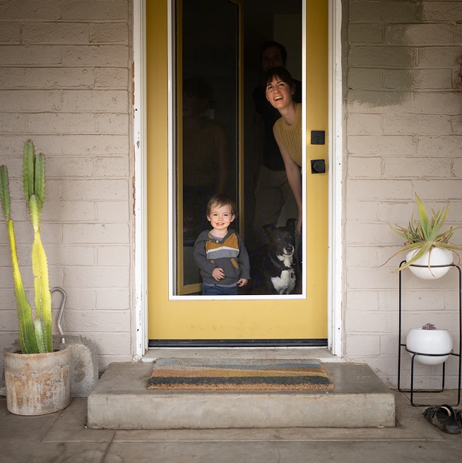 A toddler, a black dog, and the toddler's mom peer through the large window of a yellow front door.