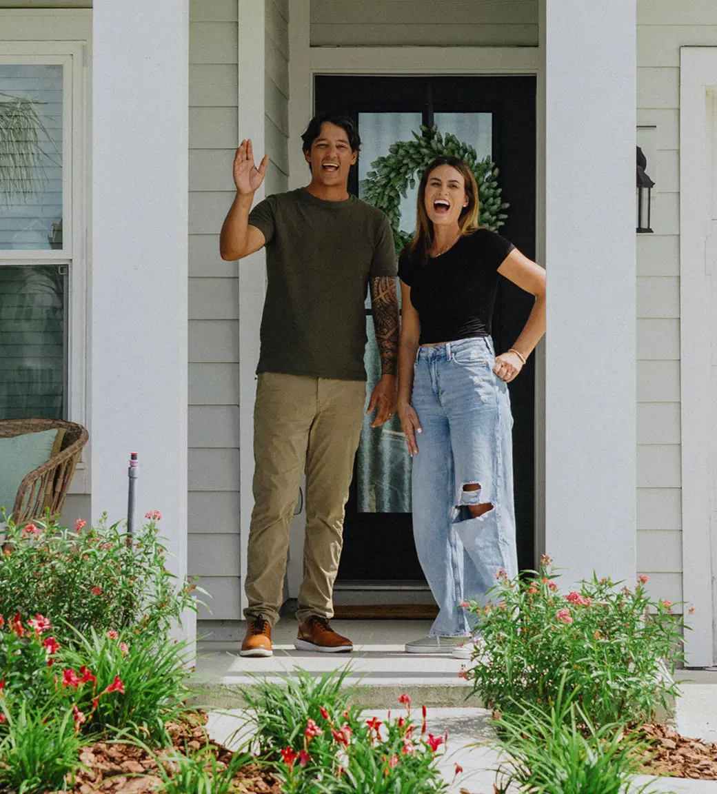 Smiling couple waving while standing on the front porch of their home.