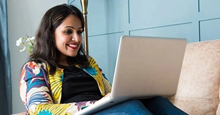 An Indian woman in a colorful jacket smiles as she researches FHA loans on her laptop.