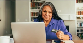 A mature woman with long silver and black hair wearing a purple shirt researches refinancing and credit on her laptop in her kitchen.